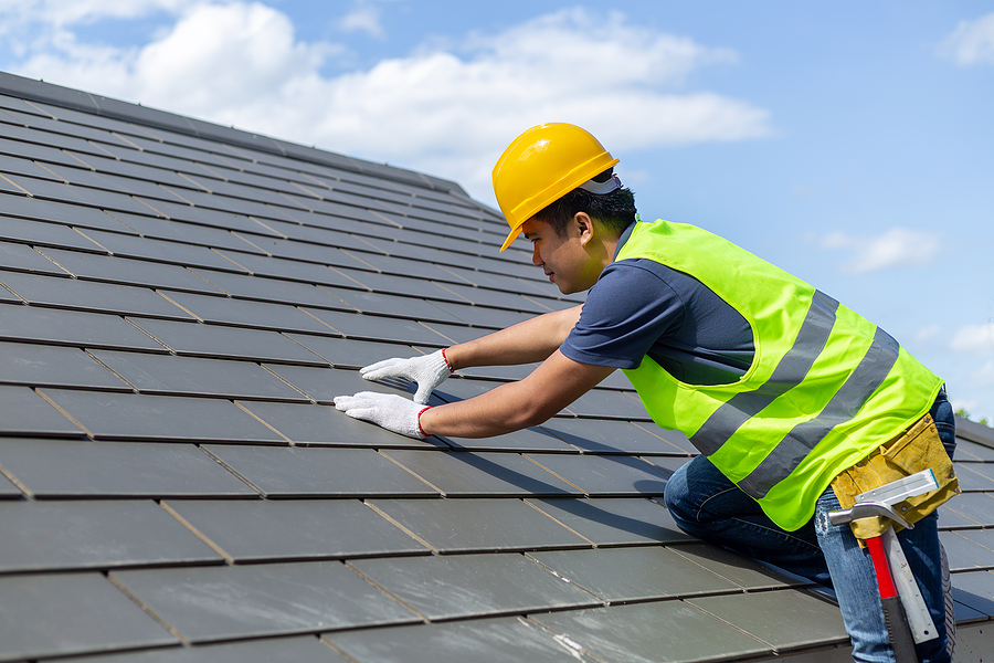 Timely Roof Repair,Roof repair, worker with white gloves replacing gray tiles or shingles on house with blue sky as background and copy space, Roofing - construction worker standing on a roof covering it with tiles.